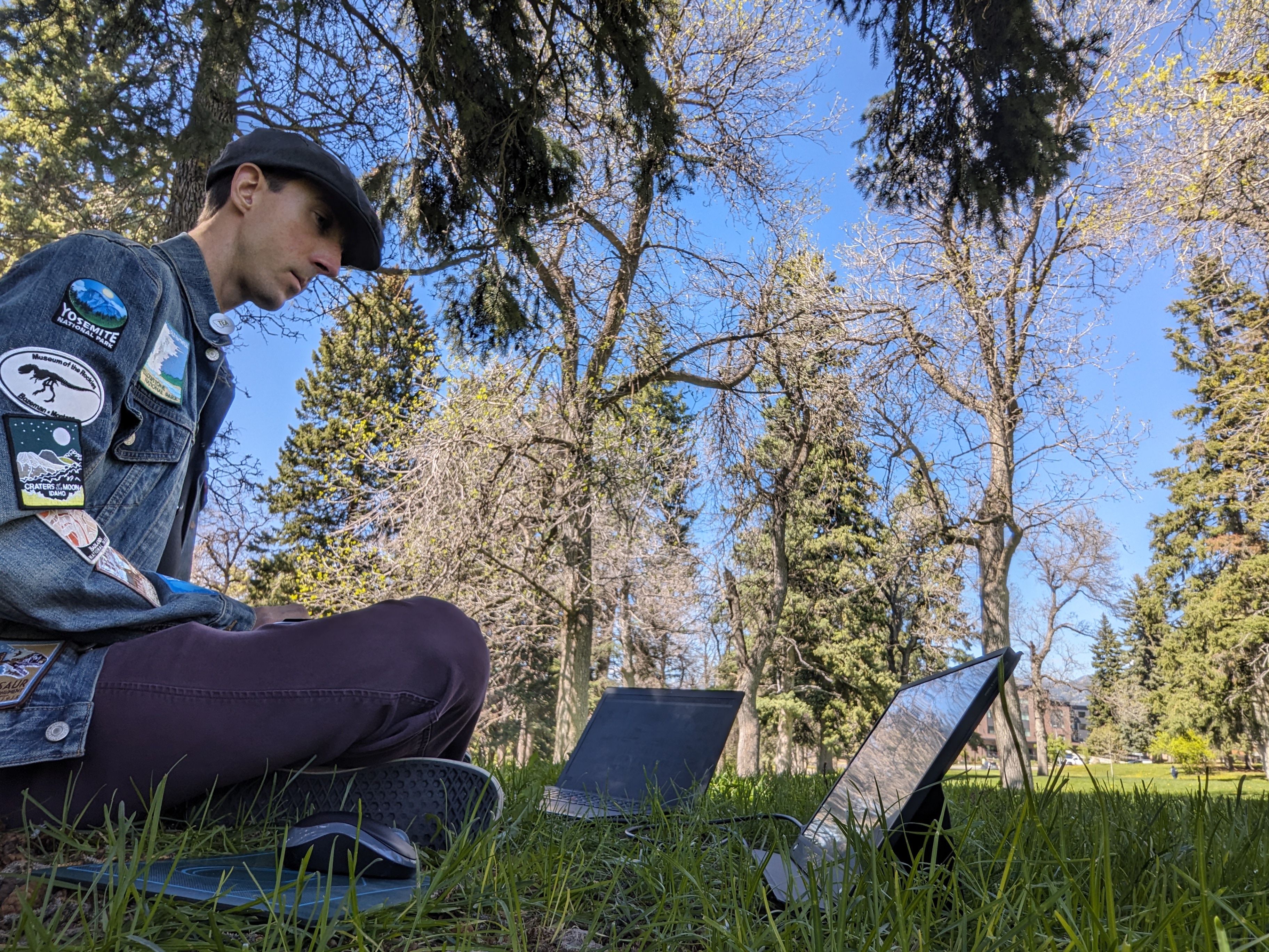 A person wearing a denim jacket sits on grass under a tree, looking at a notebook computer on the ground in front of them.